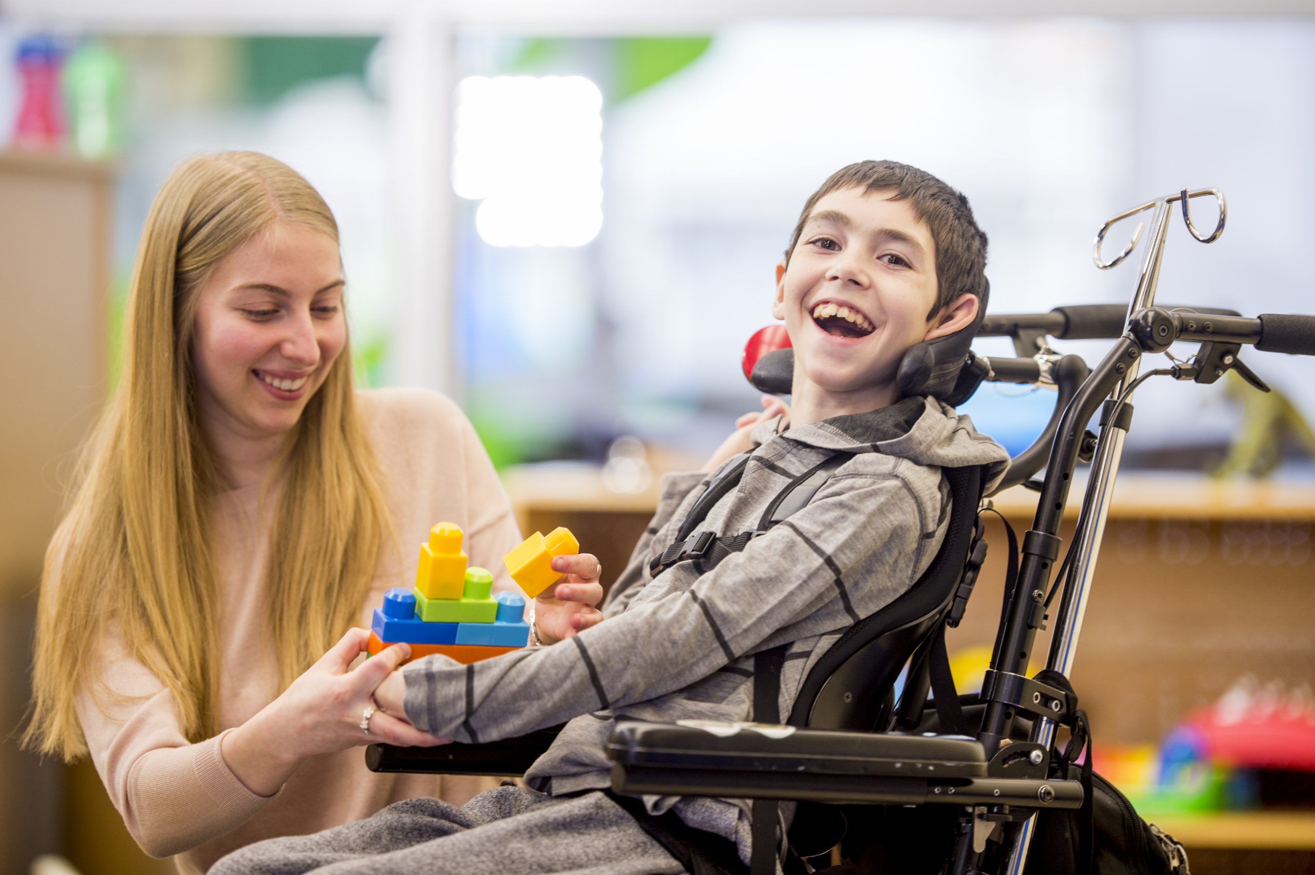 A caregiver is helping a young boy with a physical disability play with plastic block toys.